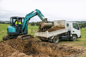 A compact teal/turquoise mini excavator with operator in cab, digging a large trench in a rural Northland paddock for a new septic tank installation. A white Mitsubishi Canter tipper truck nearby being loaded with spoil. Overcast NZ sky, green paddocks visible in background. Technician wearing a hi-vis polo shirt with royal blue collar, lime green upper half, and royal blue lower half.