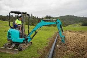 A compact teal/turquoise mini excavator with operator in cab, laying perforated drainage pipe in a trench across a rural Northland paddock for a septic disposal field. The operator wears a hi-vis polo shirt with royal blue collar, lime green upper half, and royal blue lower half. Overcast NZ sky, green paddocks and native bush in the background.