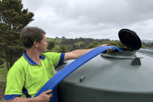 A technician in a lime green and royal blue hi-vis polo shirt holding a blue layflat hose, feeding it into the open lid on top of a large poly water tank on a rural Northland property, filling the tank. Rolling green paddocks and NZ native bush visible in the background under an overcast sky.