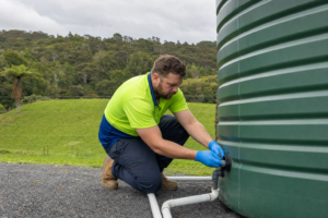 A technician in a hi-vis polo shirt with lime green upper half and royal blue collar and lower half, wearing blue PVC gloves and work boots, connecting pipework to a large rainwater tank on a rural Northland property, with green paddocks and NZ native bush visible in the background under an overcast sky